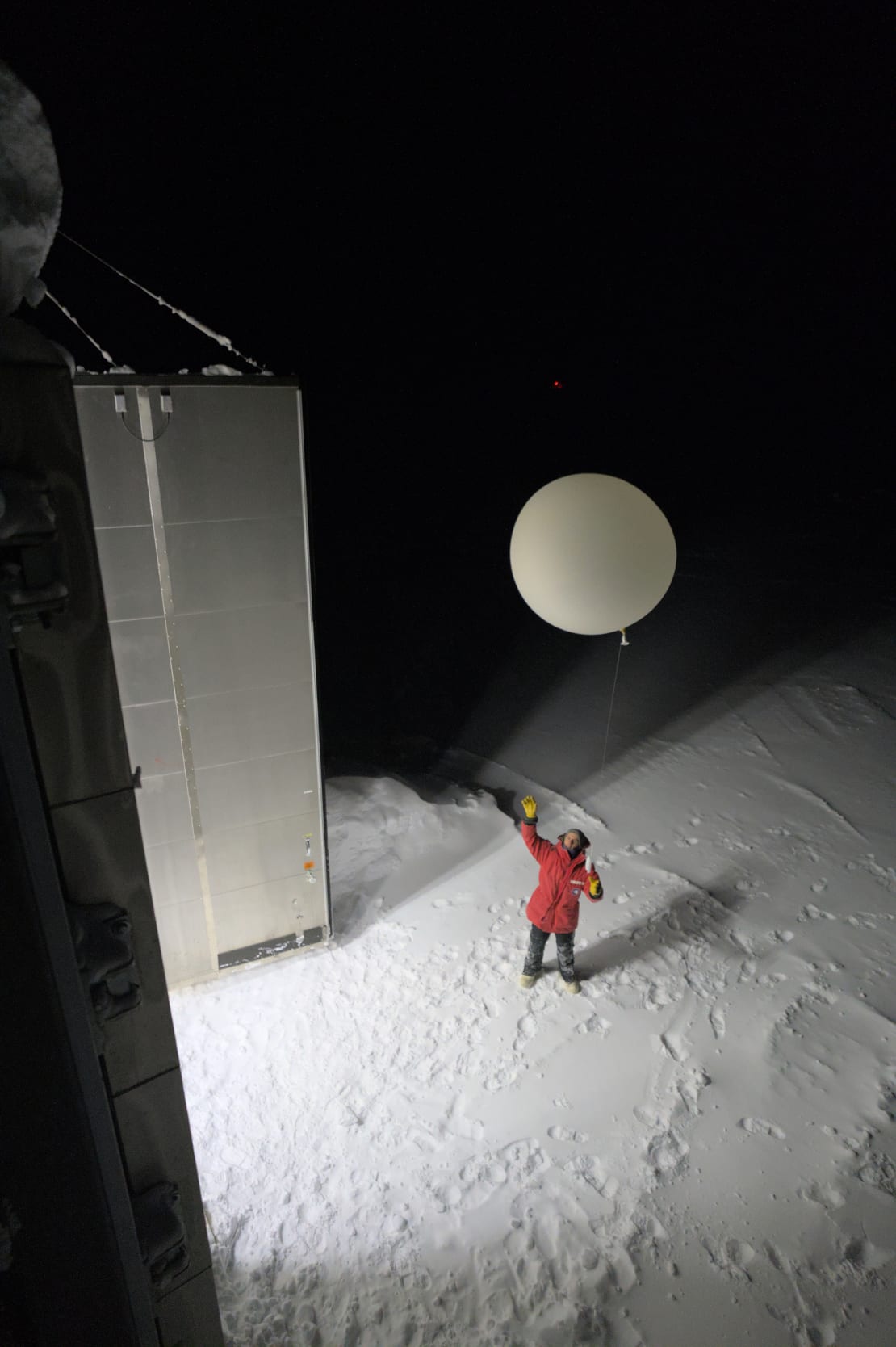 Photo from above of winterover in red parka just having launched a weather balloon outside in the dark.