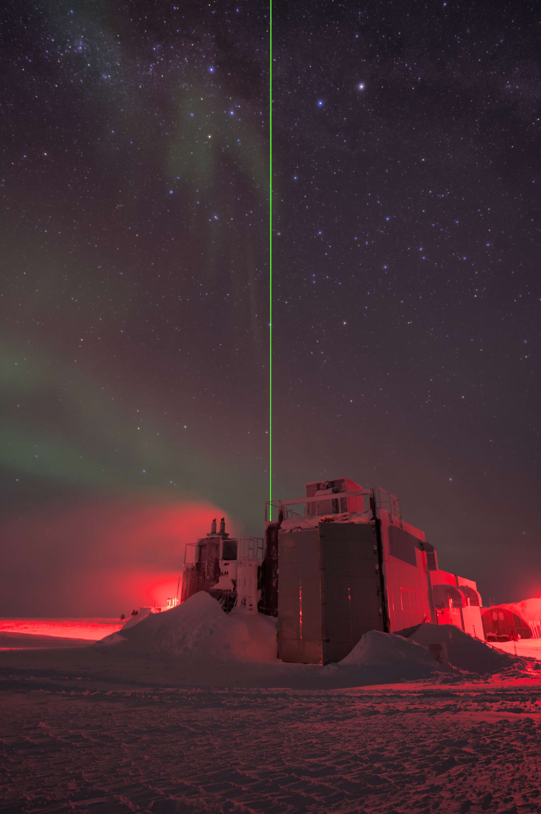 Vertical view of the cryo building at the South Pole, with a green laser shooting straight up into the starry sky from behind.