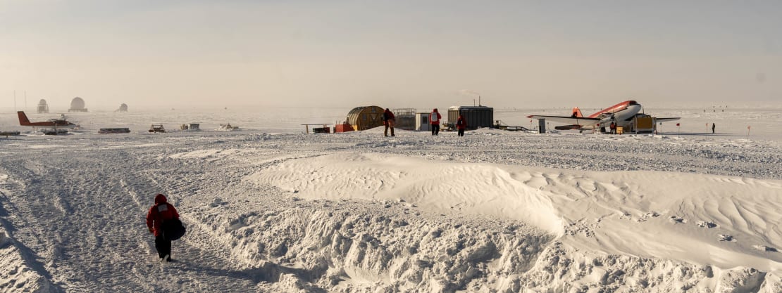 A Basler plane parked on the ice in the distance, with a lone passenger walking along worn path in snow to the station.