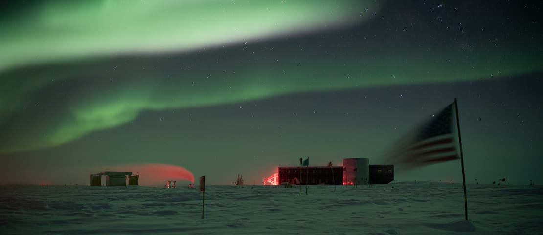 Auroras over the South Pole station in the distance, flag flapping in wind in the foreground.