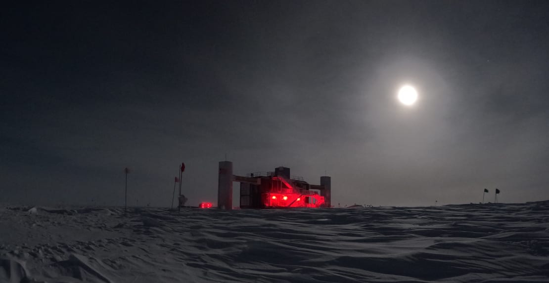 Full moon in dark sky over the IceCube Lab with its red exterior lights on.