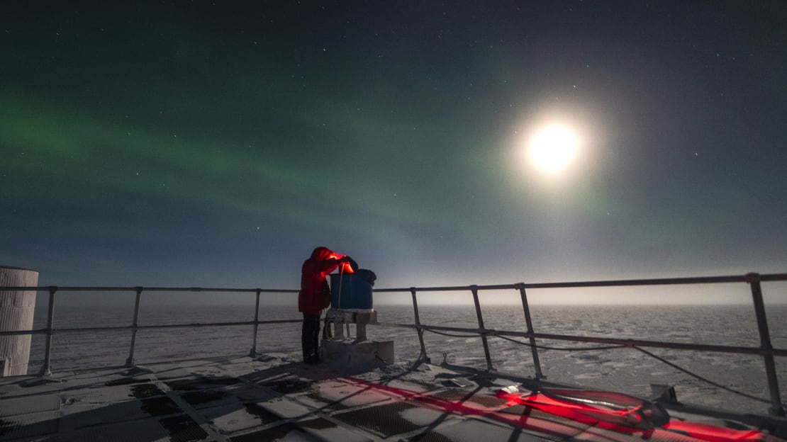 Person outdoors working on IceAct telescope, wearing red headlamp and lit from above by bright moon.