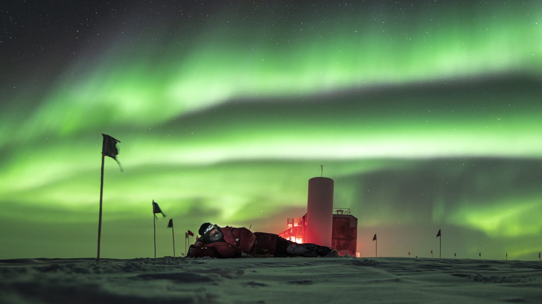 Side view of IceCube Lab with person lying on side on the ice and sky full of swirling green auroras.