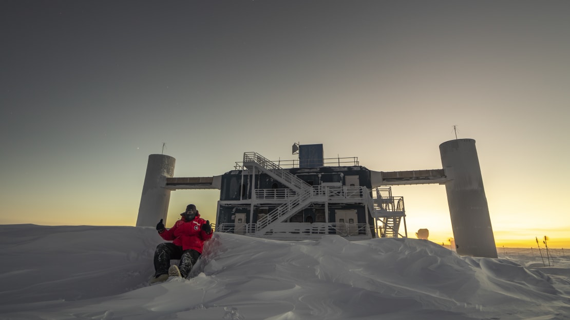 Winterover seated in snow in front of the IceCube Lab at twilight.