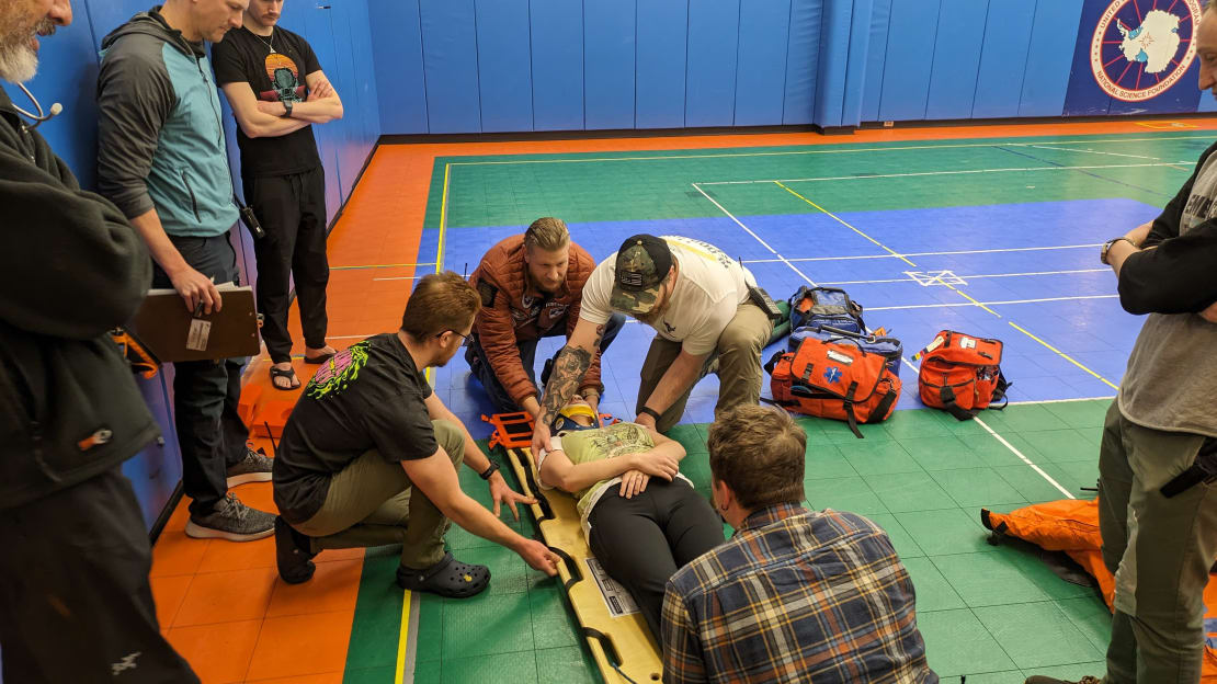 Training drill in gym at South Pole station, with several people crouched around, positioning a “patient” on a stretcher.