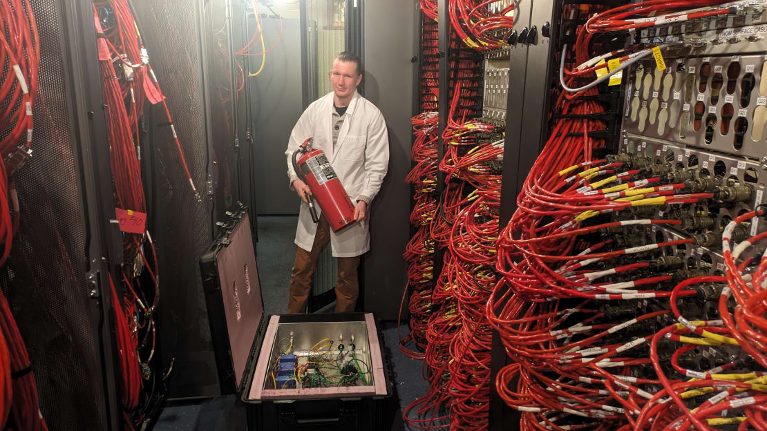 Winterover holding a fire extinguisher among racks of computer with lots of exposed orange cables.