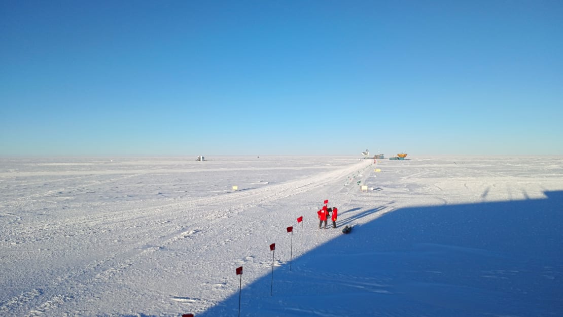 Clear blue sky on sunny day at the South Pole with bird’s-eye view of winterovers setting up a flag line, long shadow of building off to the side.