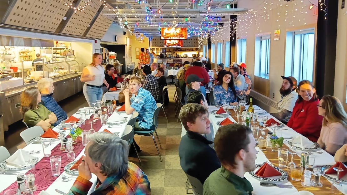 Long view down the South Pole station galley with people seated at two rows of tables set with tablecloths and dinnerware.
