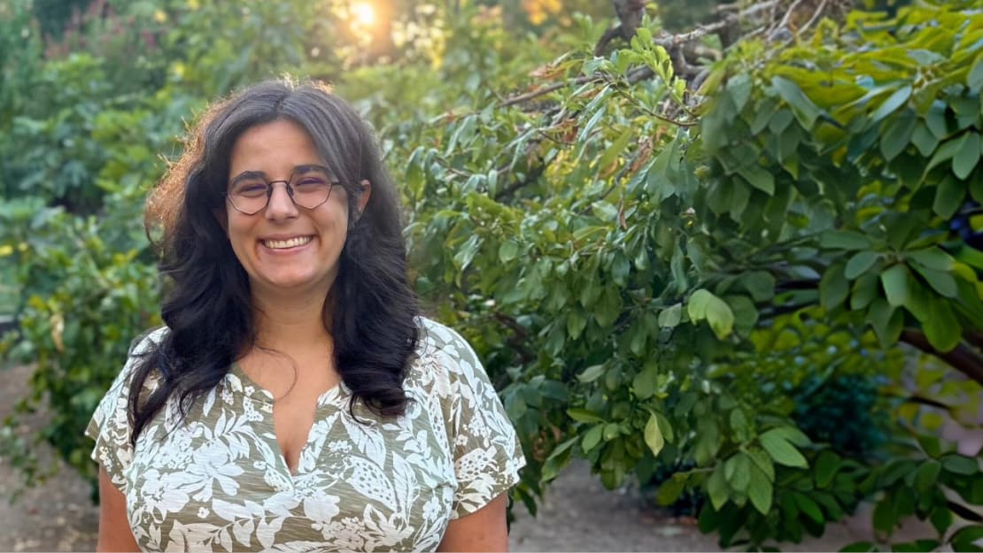 A woman with long hair and glasses smiling in front of trees.