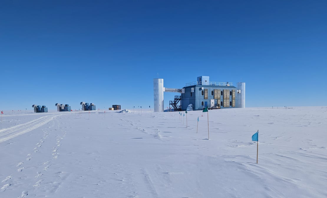 The IceCube Lab in summer under clear blue sky, with three blue power generators lined up on the ice beside it.