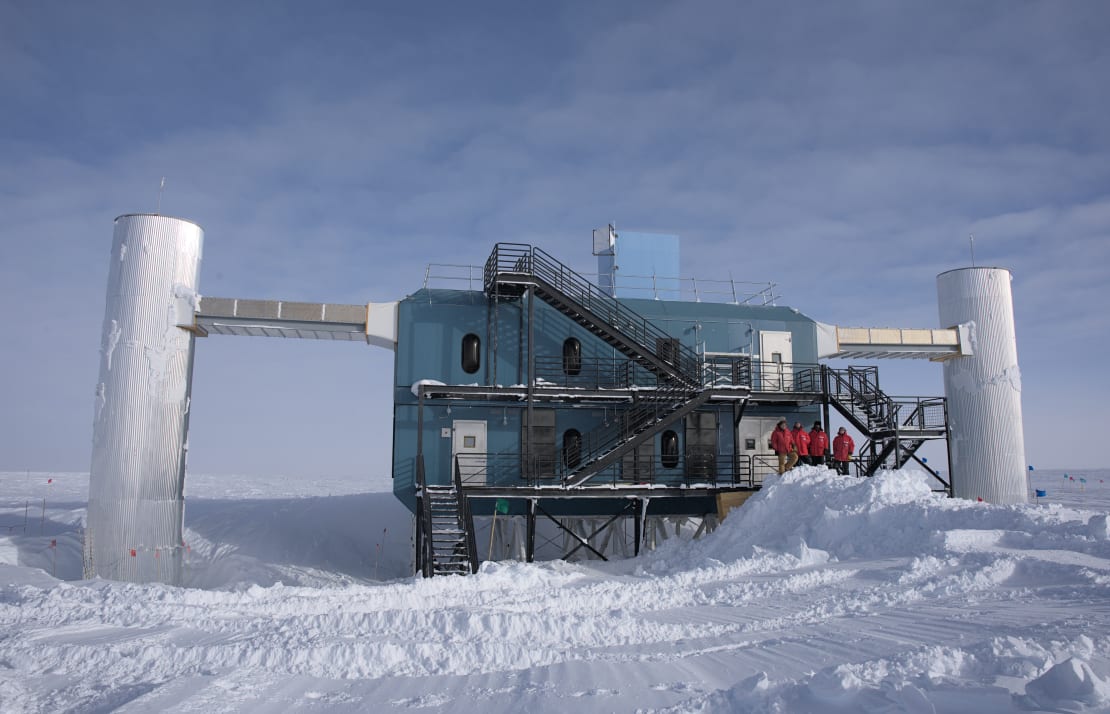 Four winterovers in red parkas standing on the platform of the IceCube Lab at a distance.