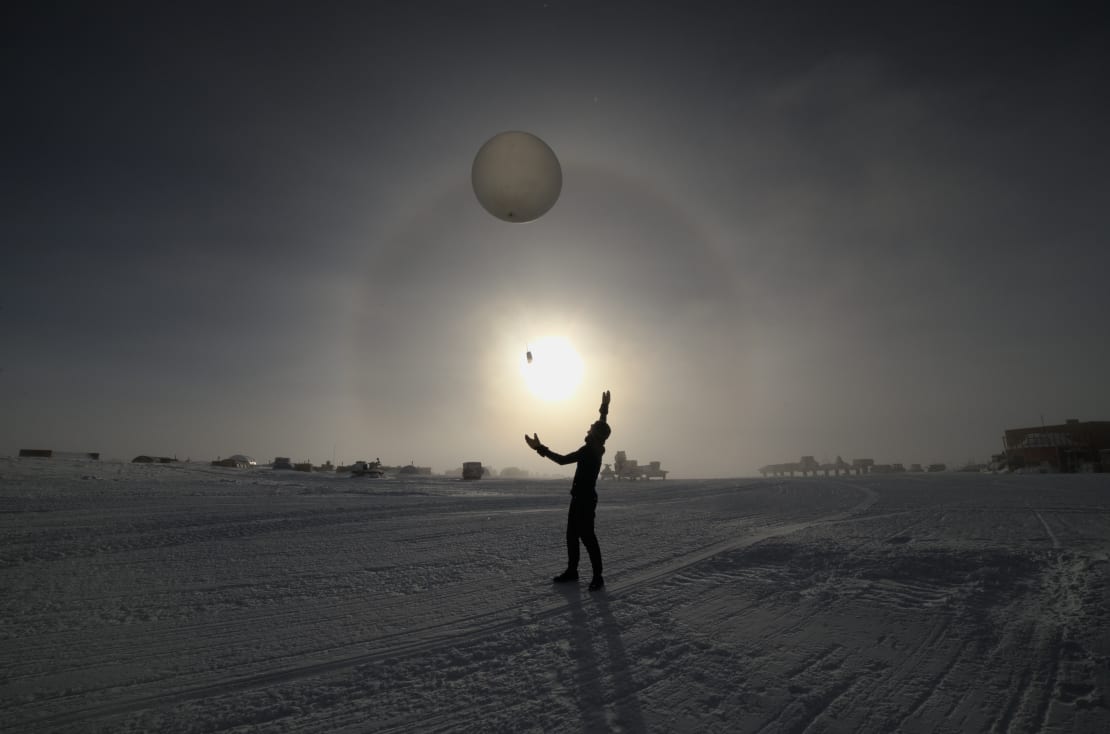 A person in shadow, having just launched a weather balloon, with the balloon a few meters above the ground and intersection the halo around the low sun in the sky.