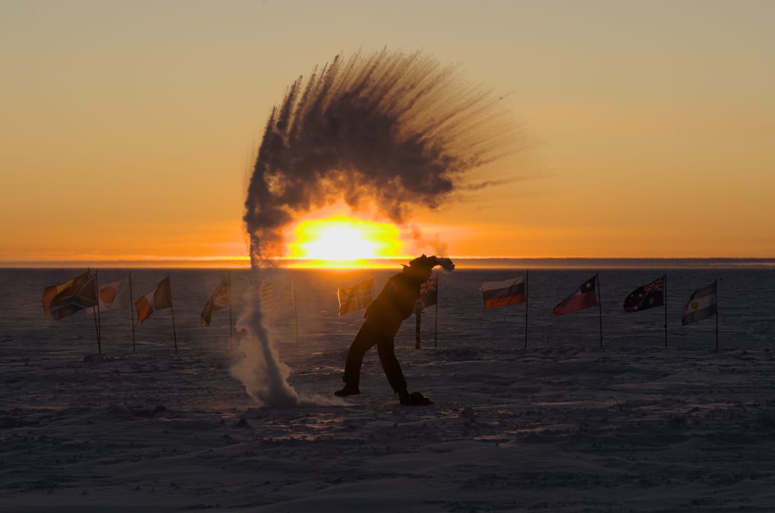 A person in shadow as they are throwing boiling water into the air, creating an arc of ice crystals, in front of the sunset at the ceremonial Pole.