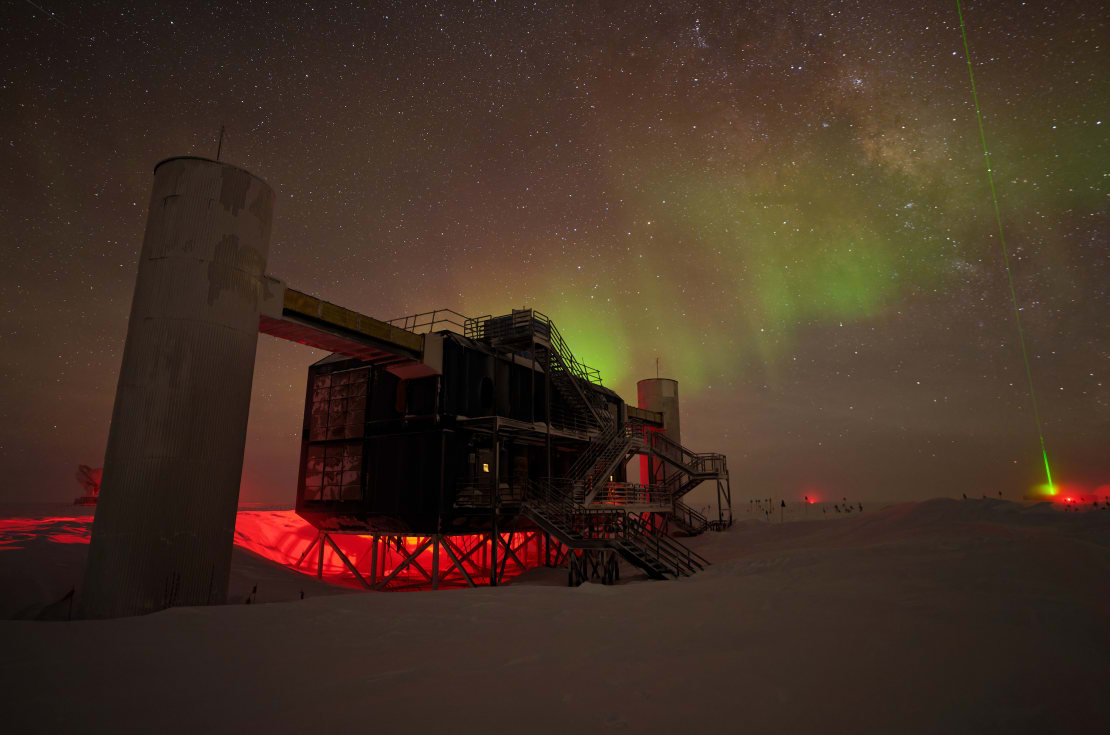 The IceCube Lab in winter shadowed by bright auroras in the background.