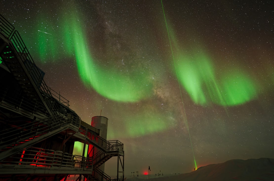 Very curvy green auroras scrawled across the sky, IceCube Lab close up in lower left corner.