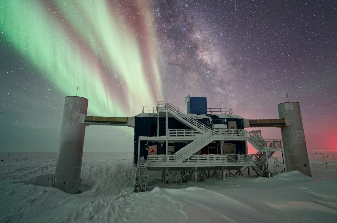 The IceCube Lab with a pseudo-split-screen background of Milky Way and stars off to the right and auroras off to the left.