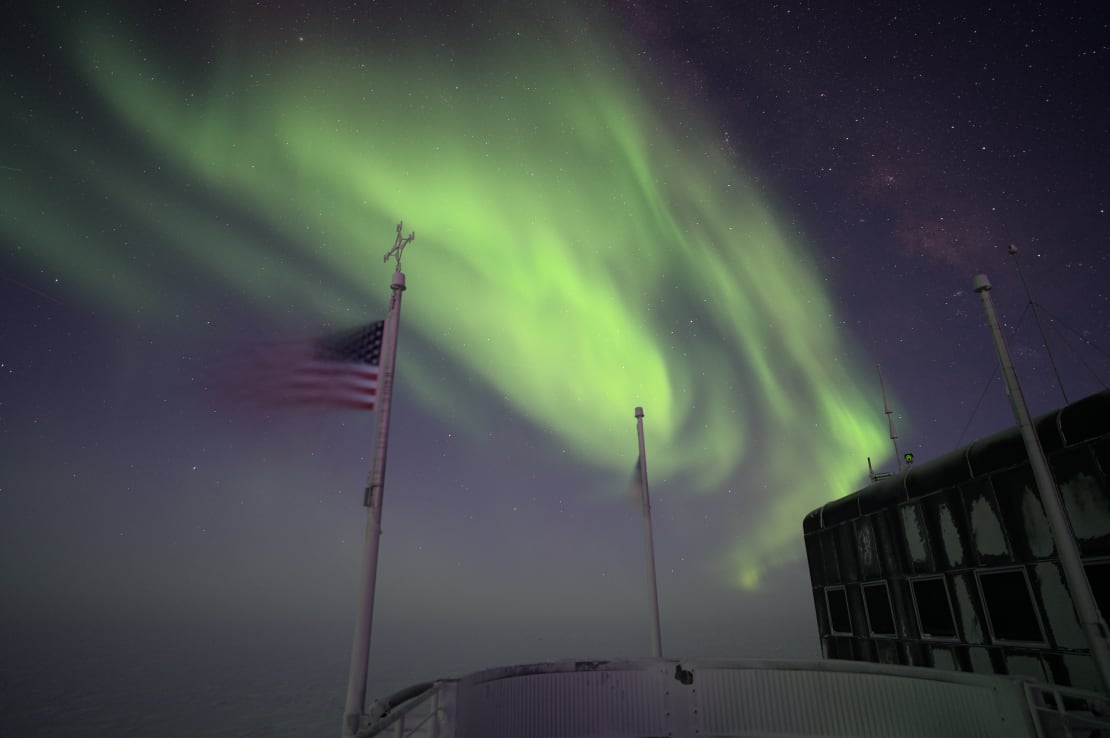 Bright green blob of auroras over top corner of South Pole station, with US flag whipping in the wind.