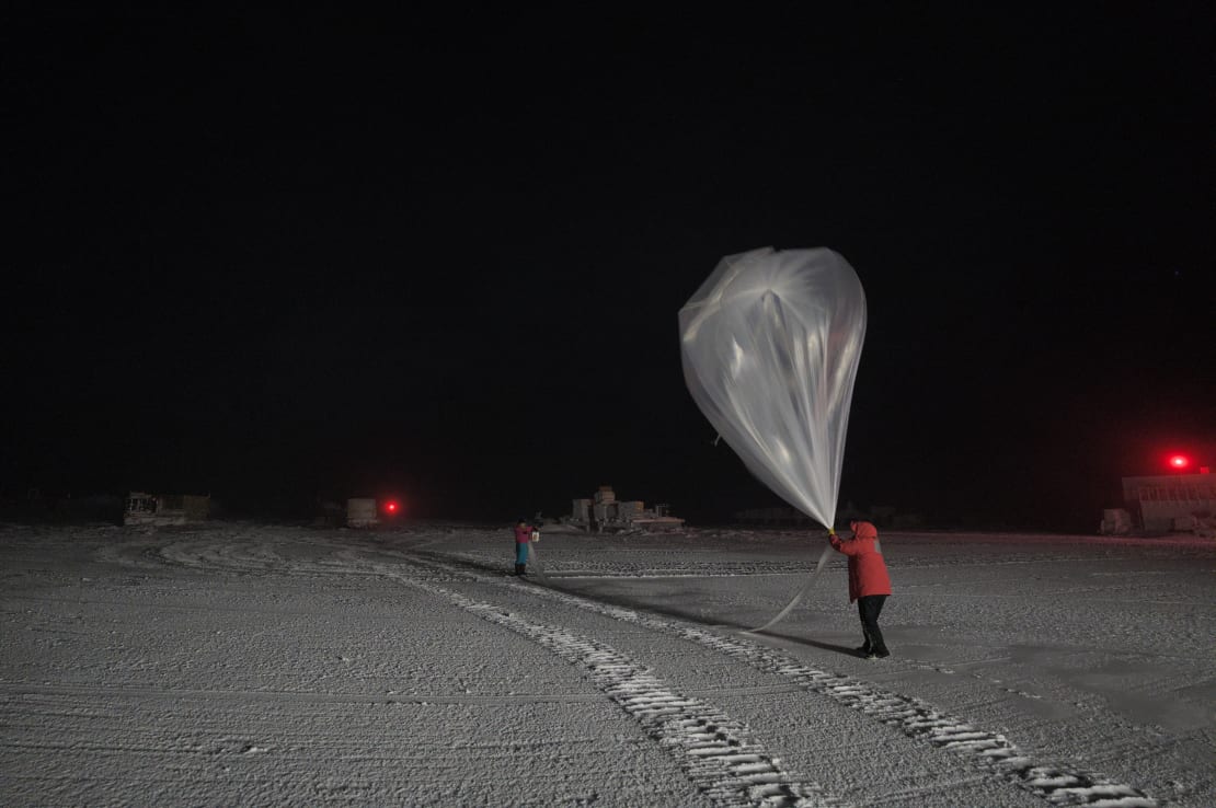 Winterover in red parka, against dark black sky, holding weather balloon for launching.