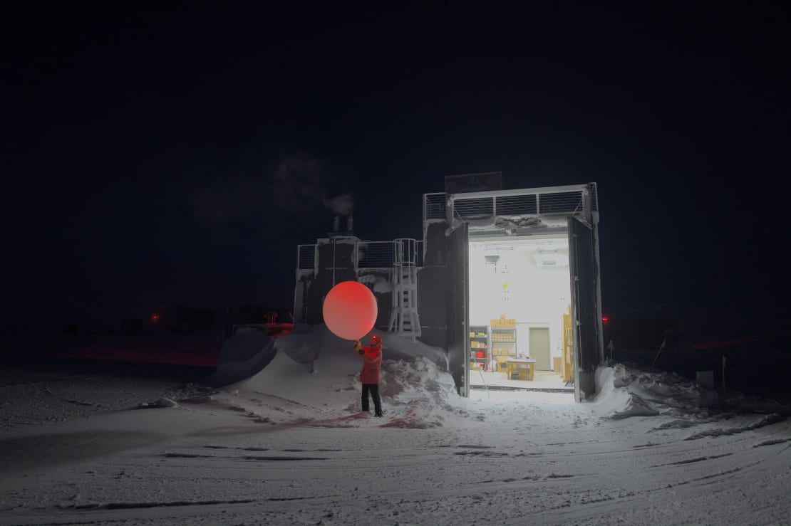 Winterover launching weather balloon in dark surroundings, only light available from doorway of building.