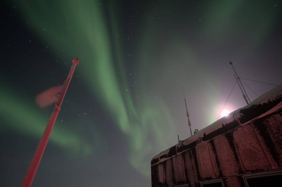 Auroras overhead and bright moon peeking over top of South Pole station.