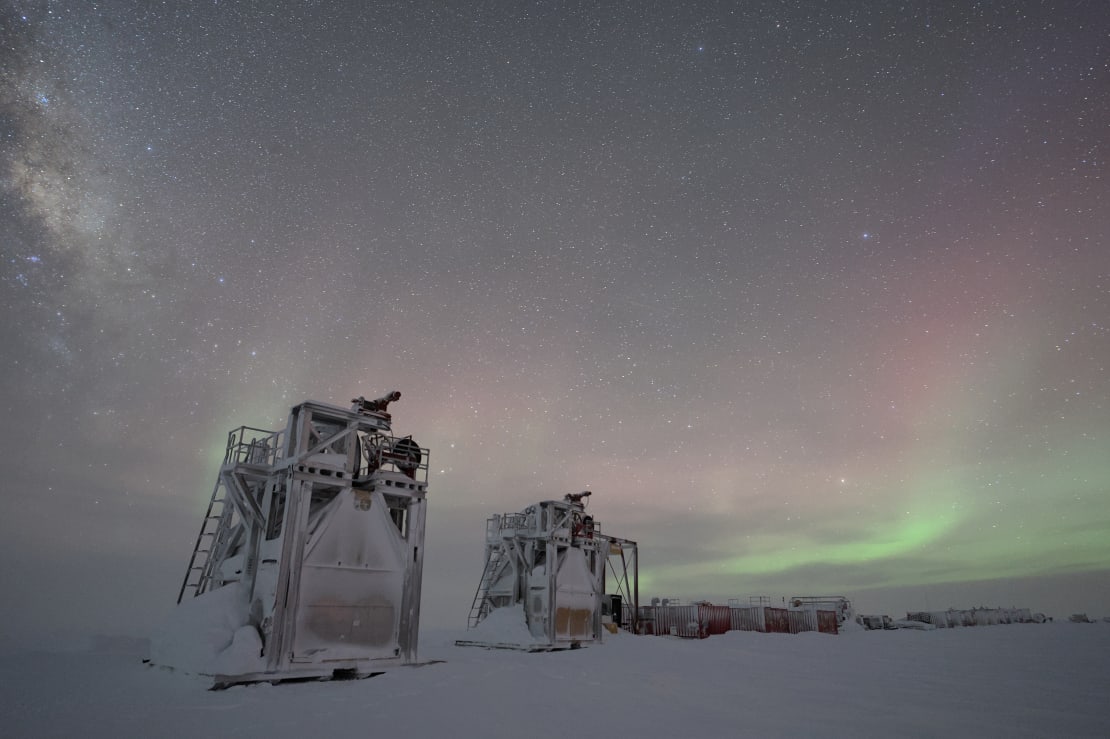 Wintry scene of two frosted and snow-covered IceCube drill towers with some faint auroras in the background.