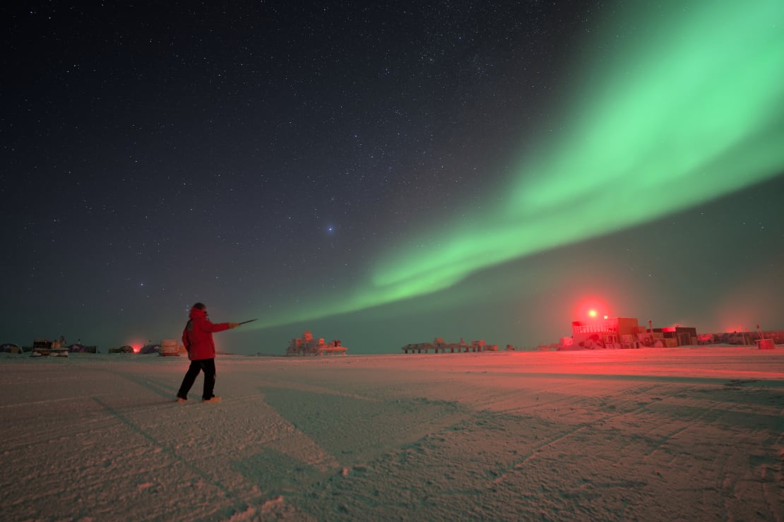 A bright streak of aurora in the sky seeming to shoot forth from a winterover’s forward-stretched arm.