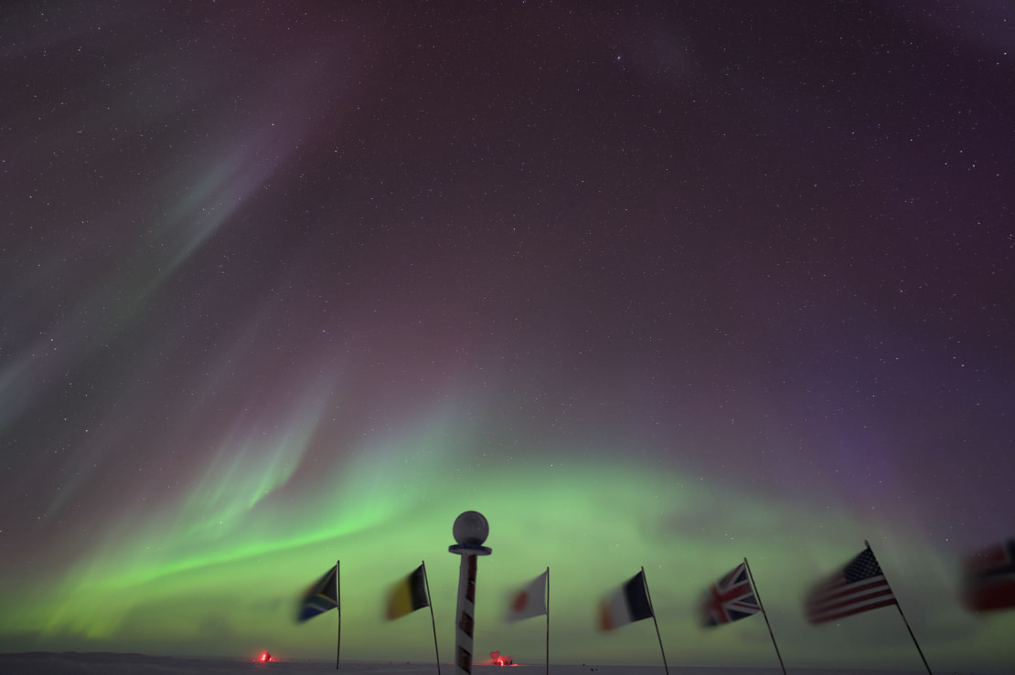 Marker and flags at ceremonial South Pole shown in foreground, bright green auroras in background, low in dark night sky.