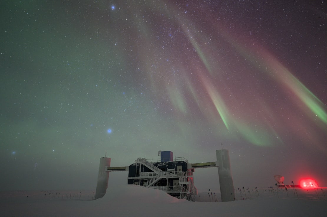 The IceCube Lab in winter, snowdrift in front, auroras overhead.