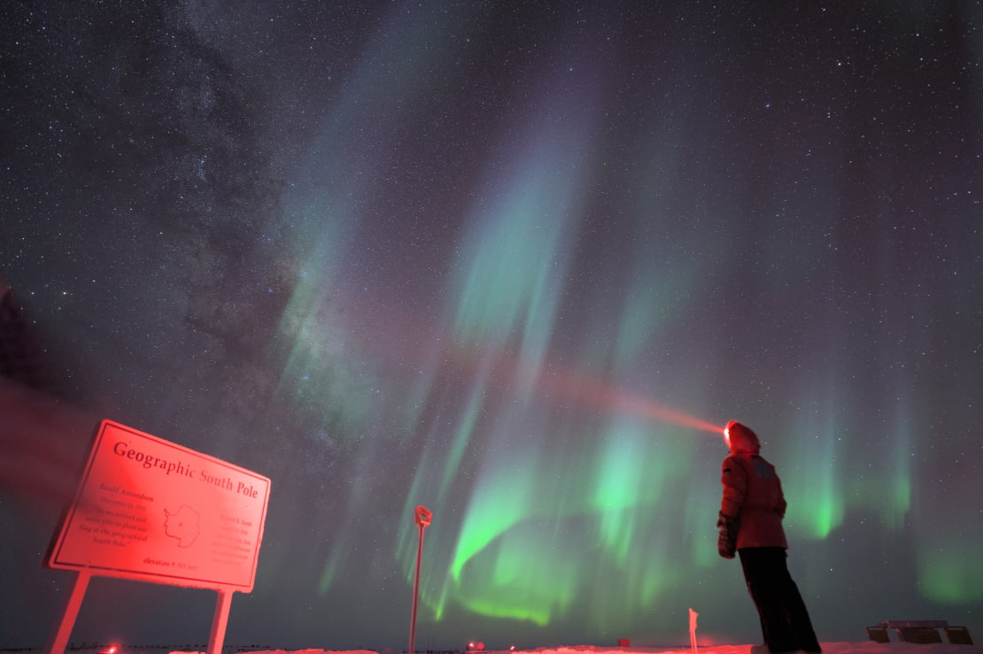 Winterover standing with red headlamp facing the geographic South Pole sign, with sky full of green and blue swirling auroras.