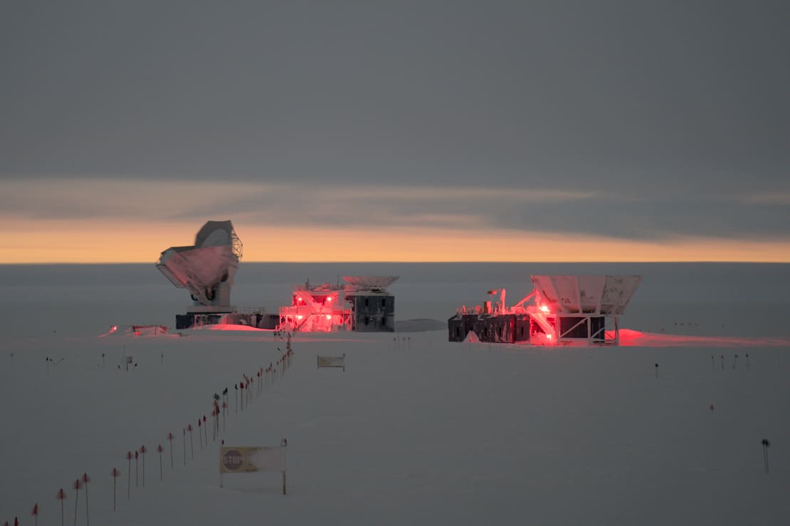 Orange glow along horizon with South Pole telescopes in distance still lit in red lights during winter-summer twilight.