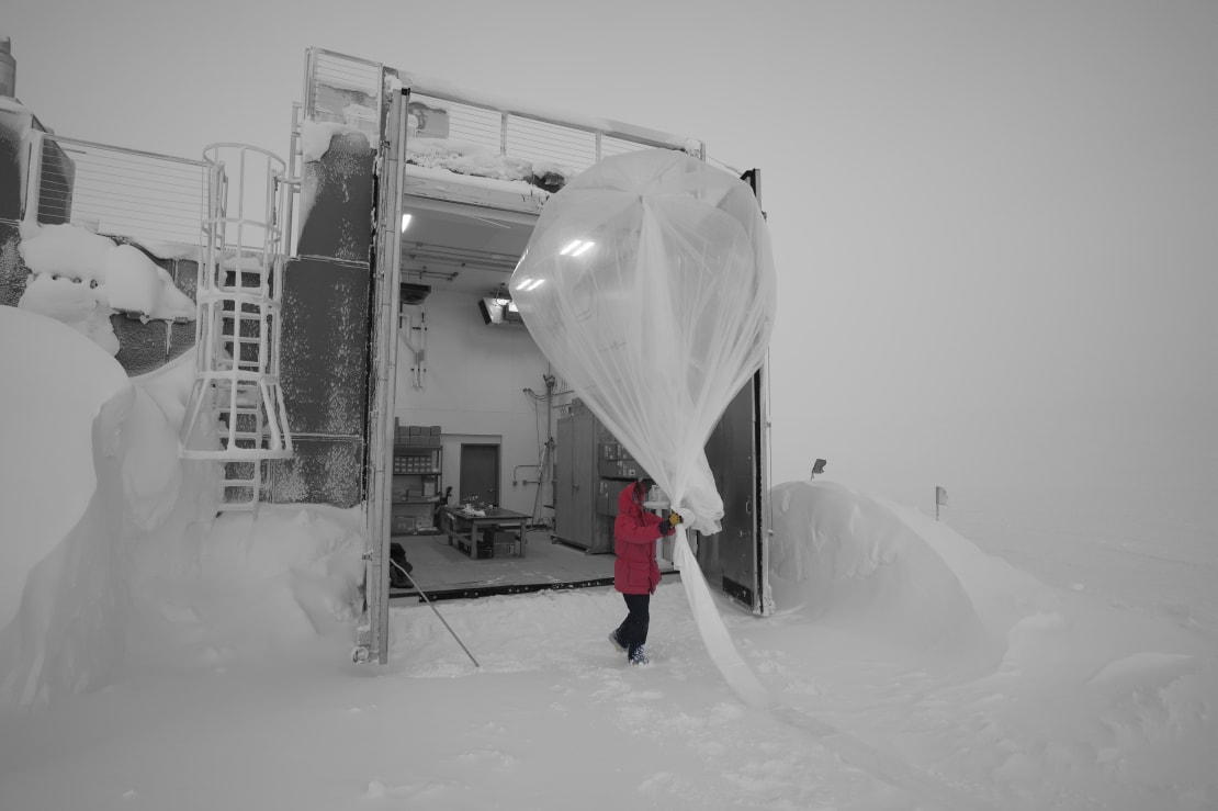 Winterover in red parka about to launch weather balloon on very cloudy day, can’t distinguish white sky from white ground.