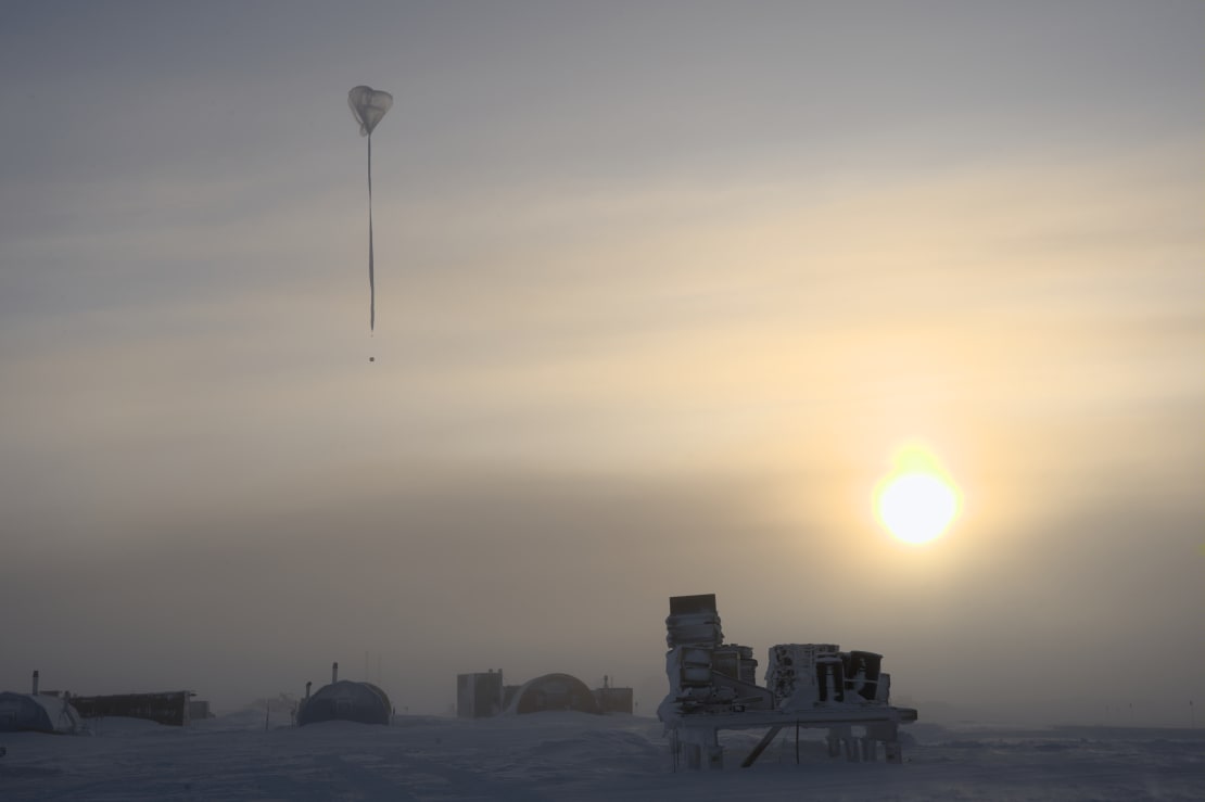 Weather balloon shown in its ascent against cloudy sky but with low bright sun showing through.