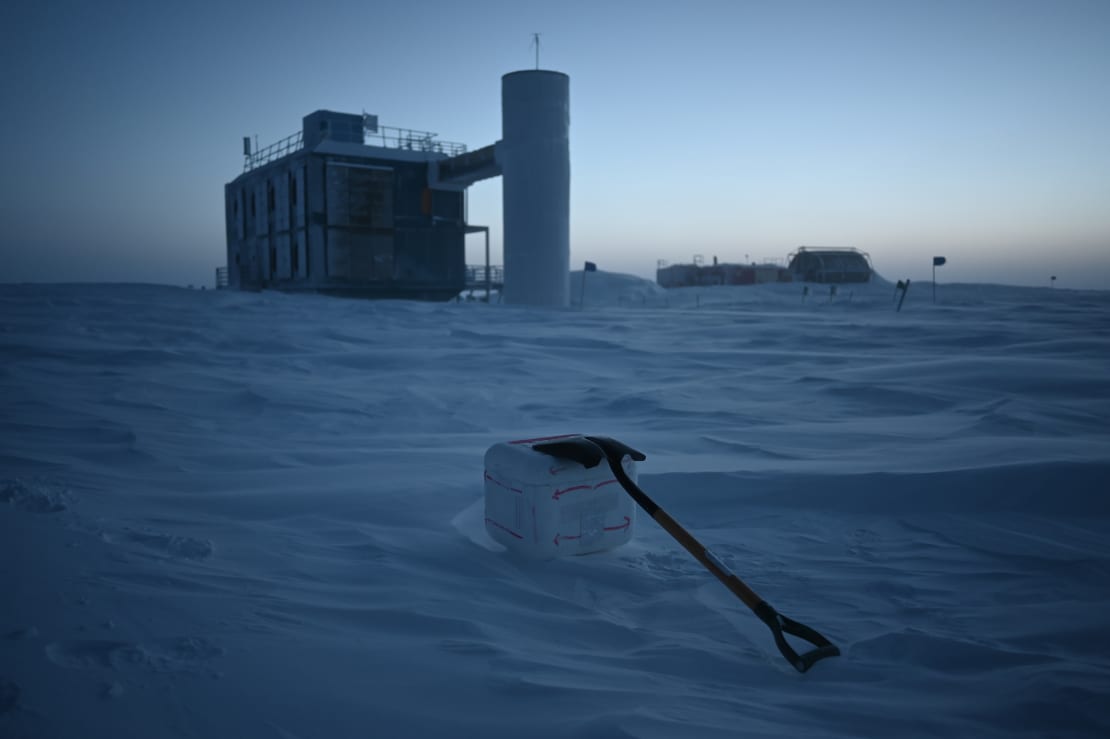 A shovel lying on top of and holding down a camera box setup out on the ice, with IceCube Lab in background.