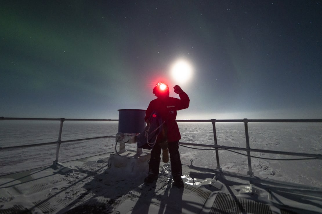 Person wearing red headlamp facing camera and waving, standing next to IceAct telescope, with bright moon just above their head.