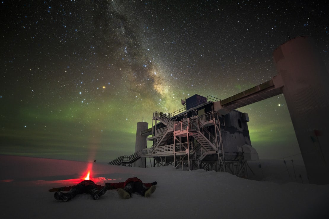 Two winterovers lying on ground outside the IceCube Lab, looking up into sky with stars and Milky Way.