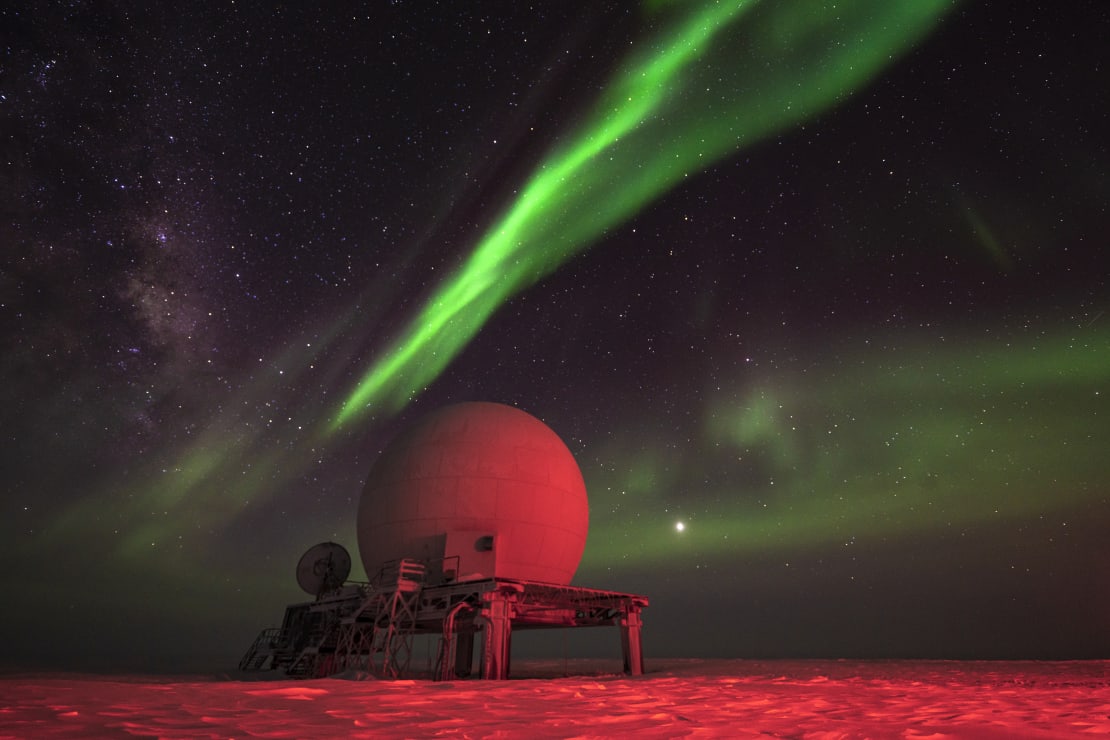 Satellite dome lit in red light, with streak of bright green aurora overhead.