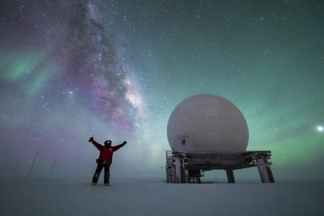 Sky full of auroras and Milky Way, person standing with arms up near a satellite dome.