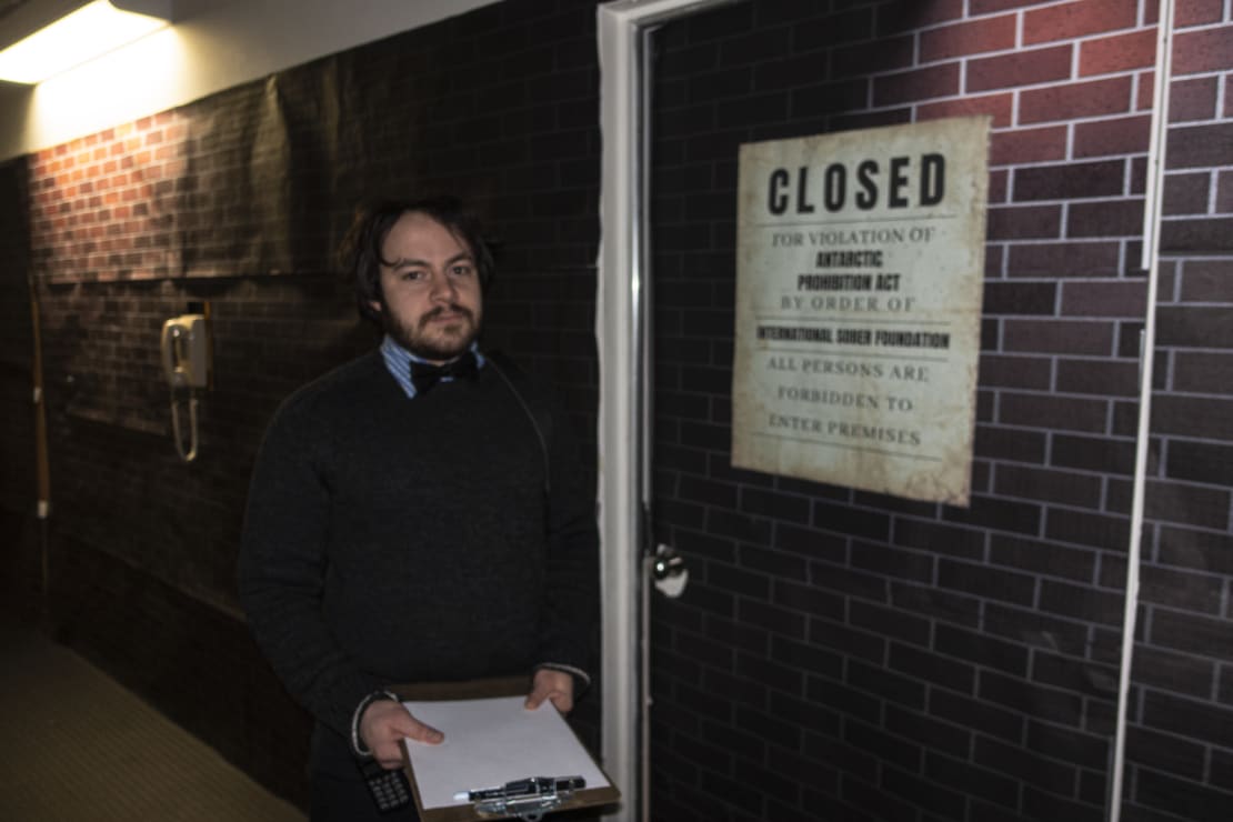 Winterover Josh standing with clipboard, as midwinter secret bar bouncer.