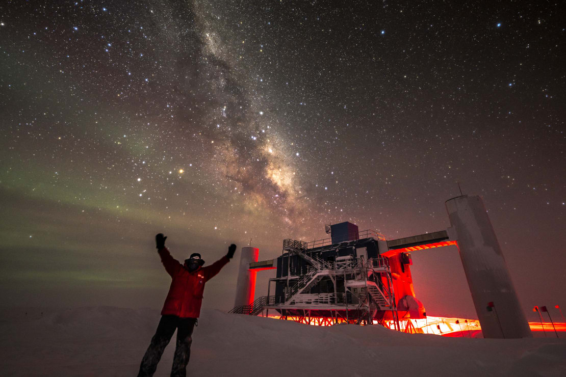 Person in red parka in front of IceCube Lab, stars and Milky Way overhead.