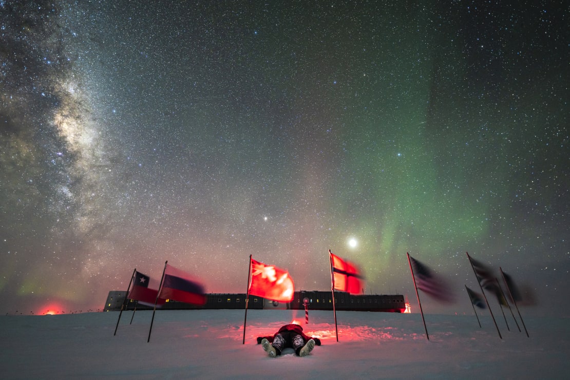 Winterover Martin lying on ground among flags at ceremonial Pole under starry skies, with view of station behind.