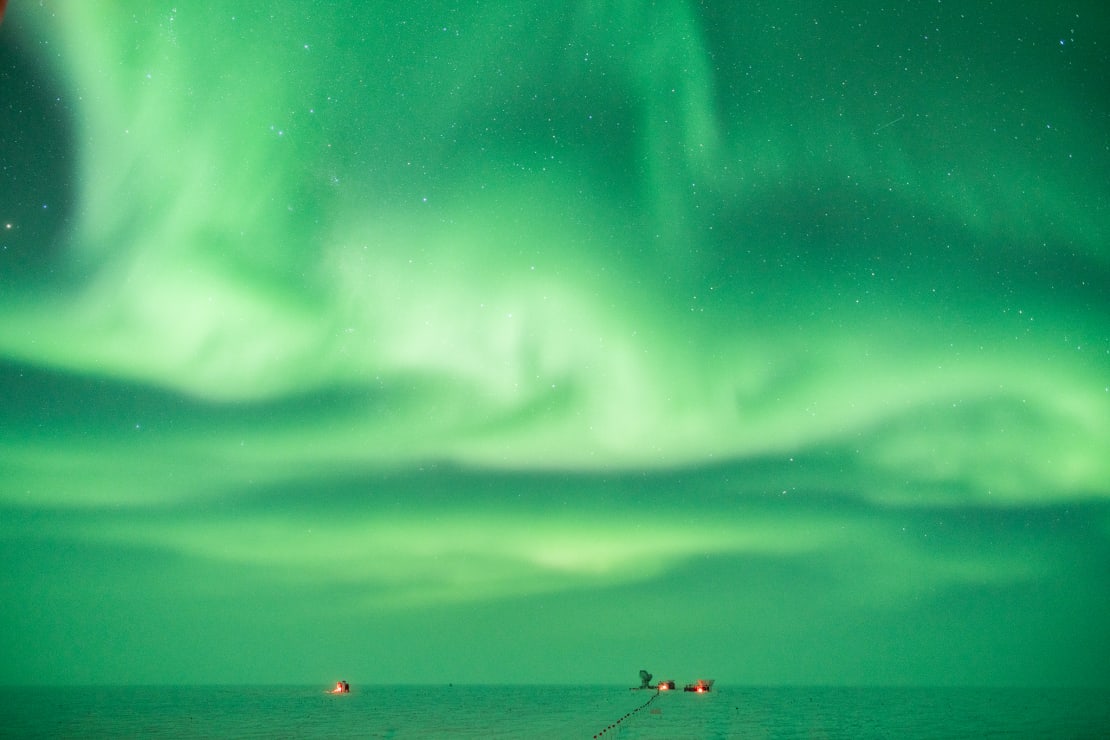 Long exposure of sky filled with green auroras, with South Pole Dark Sector buildings very small on the horizon.