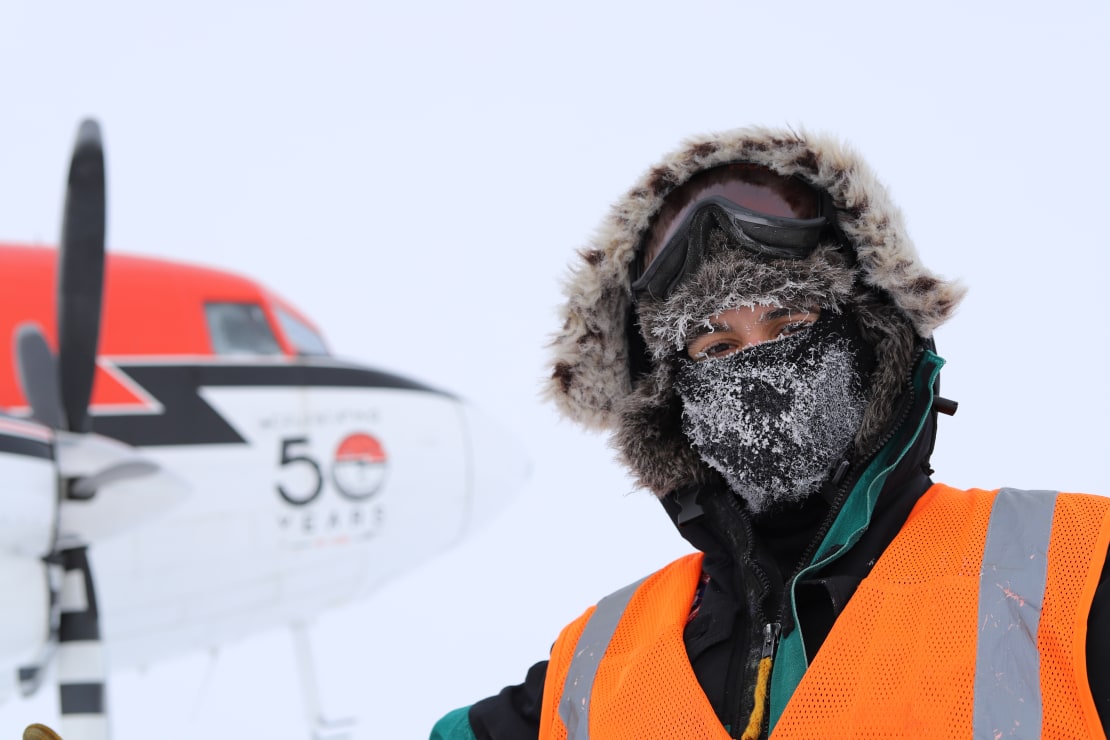 Close-up of person in parka and frosted-over balaclava out on the South Pole ice, front of small aircraft blurred in background.