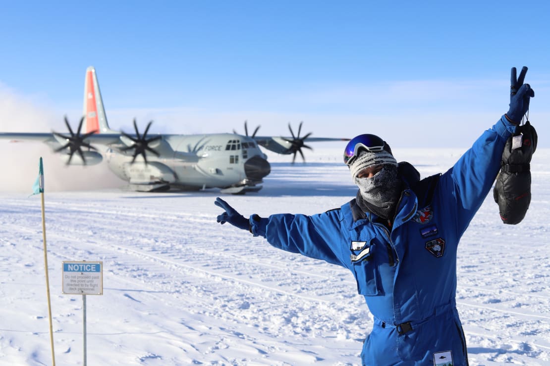 Winterover facing the camera with arms up in front of an LC-130 on the South Pole skiway.