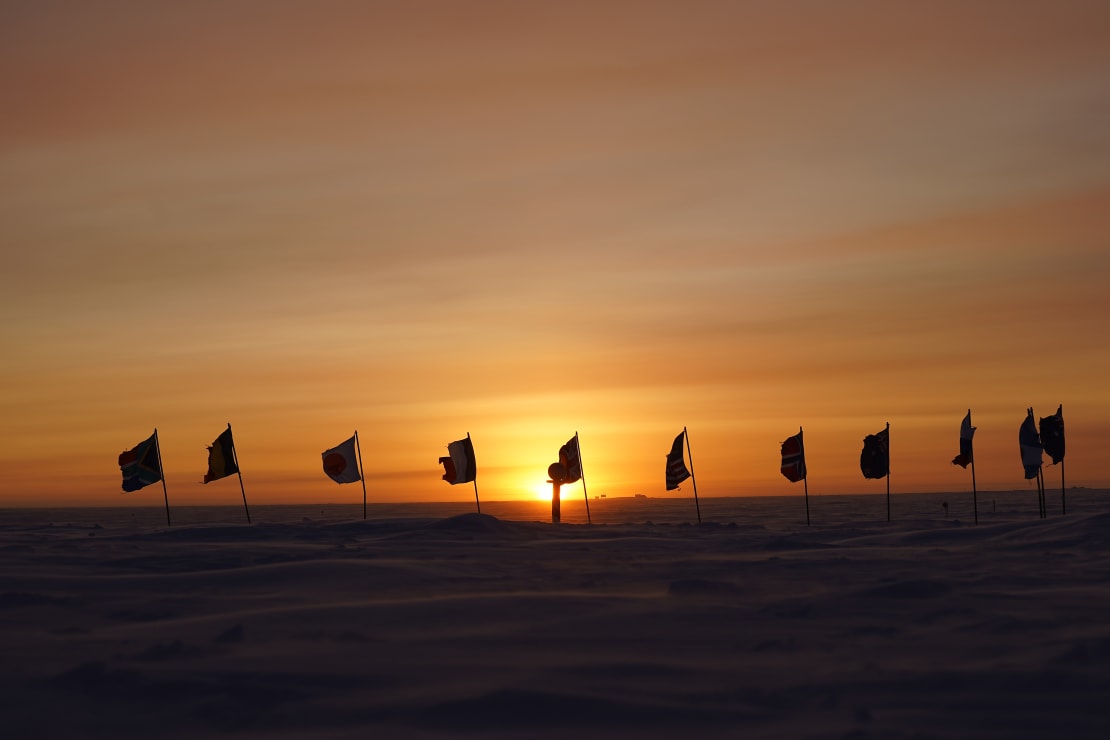 Bright orange filling the sky along horizon behind the tattered flags of ceremonial South Pole in shadow.