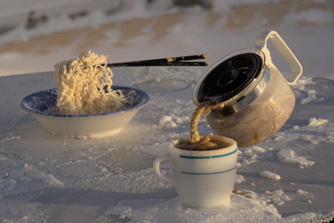 Close-up of frozen food, chopsticks suspended in clump of noodles pulled up from bowl and coffee pot suspended by frozen stream of coffee poured into cup.