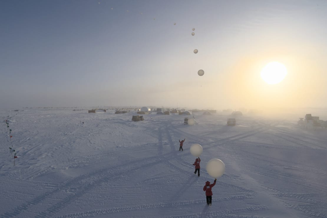 Composite image of winterover in red parka launching a weather balloon under bright sun.