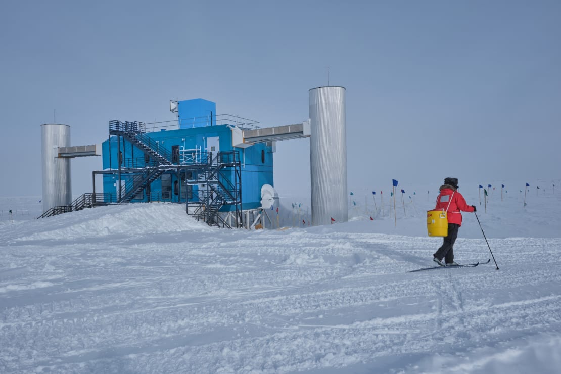 A winterover in red parka on cross country skis with IceCube Lab in background.