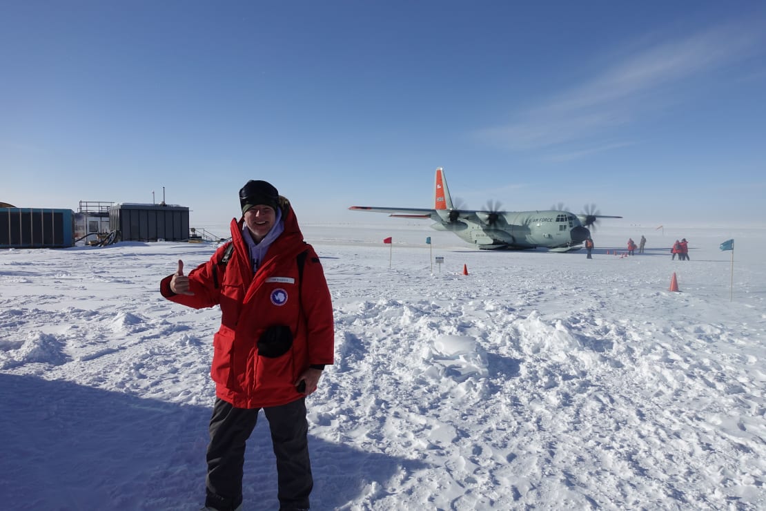 A departing winterover in red parka out on the ice giving a thumbs up, plane parked in background.