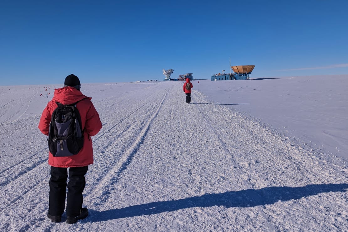 A winterover in red parka shown from behind as they film another winterover in red parka who is walking away from them toward the South Pole Telescope in the distance.