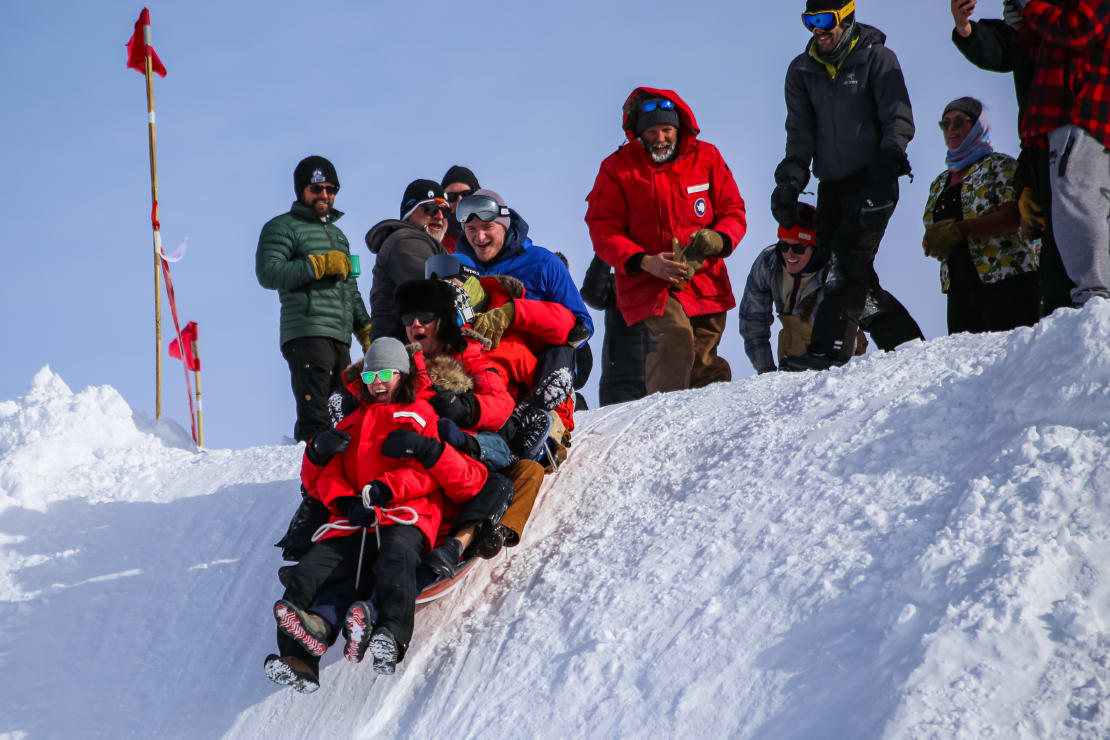 Several winterovers packed together on small sled, just starting to go down steep snowbank.